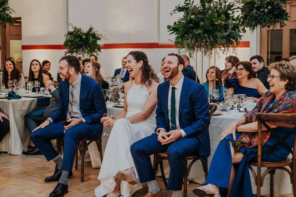 Bride and Groom laughing during toasts, seated at their Eastern Market reception surrounded by joyful family and friends.