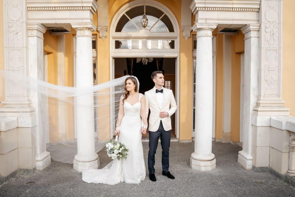 Bride and Groom stand outside of a formal building, looking away from each other. Bride's long veil is being blown horizontally to the edge of the frame.
Photography @joyphoto