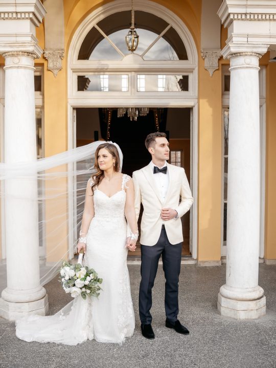 Bride and Groom stand outside of a formal building, looking away from each other. Bride's long veil is being blown horizontally to the edge of the frame. Photography @joyphoto